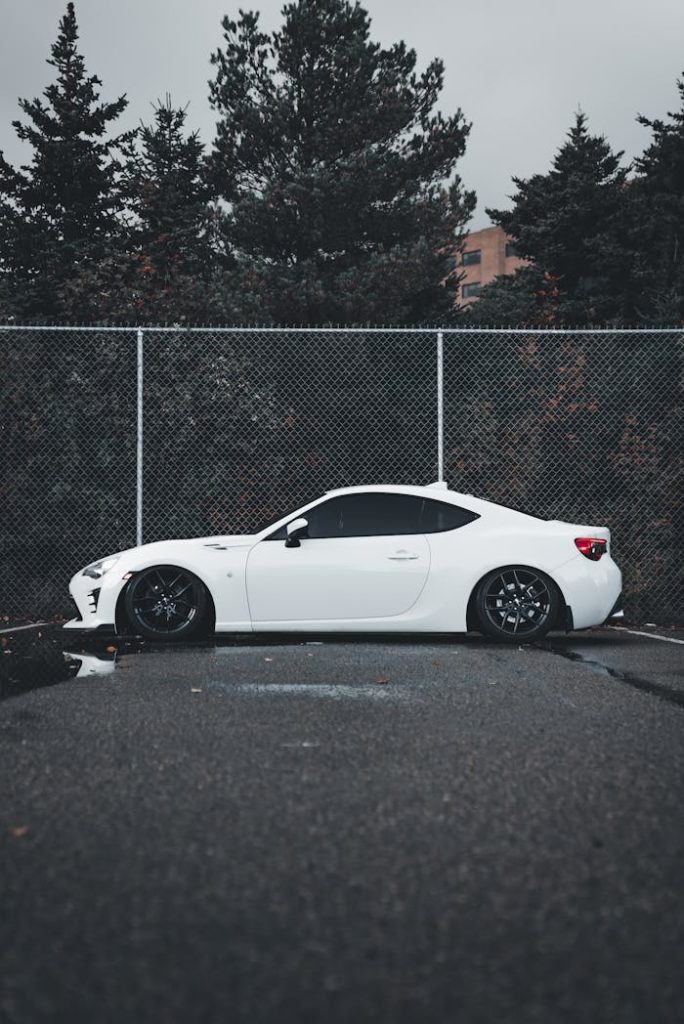 A stylish white sports car parked outdoors beside a chain-link fence on an overcast day.